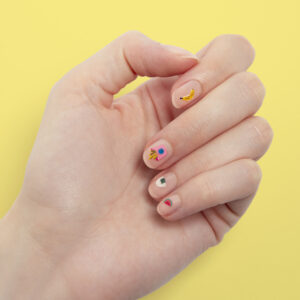 Main présentant une manucure colorée avec des motifs de banane, pastèque et sucette sur fond jaune
