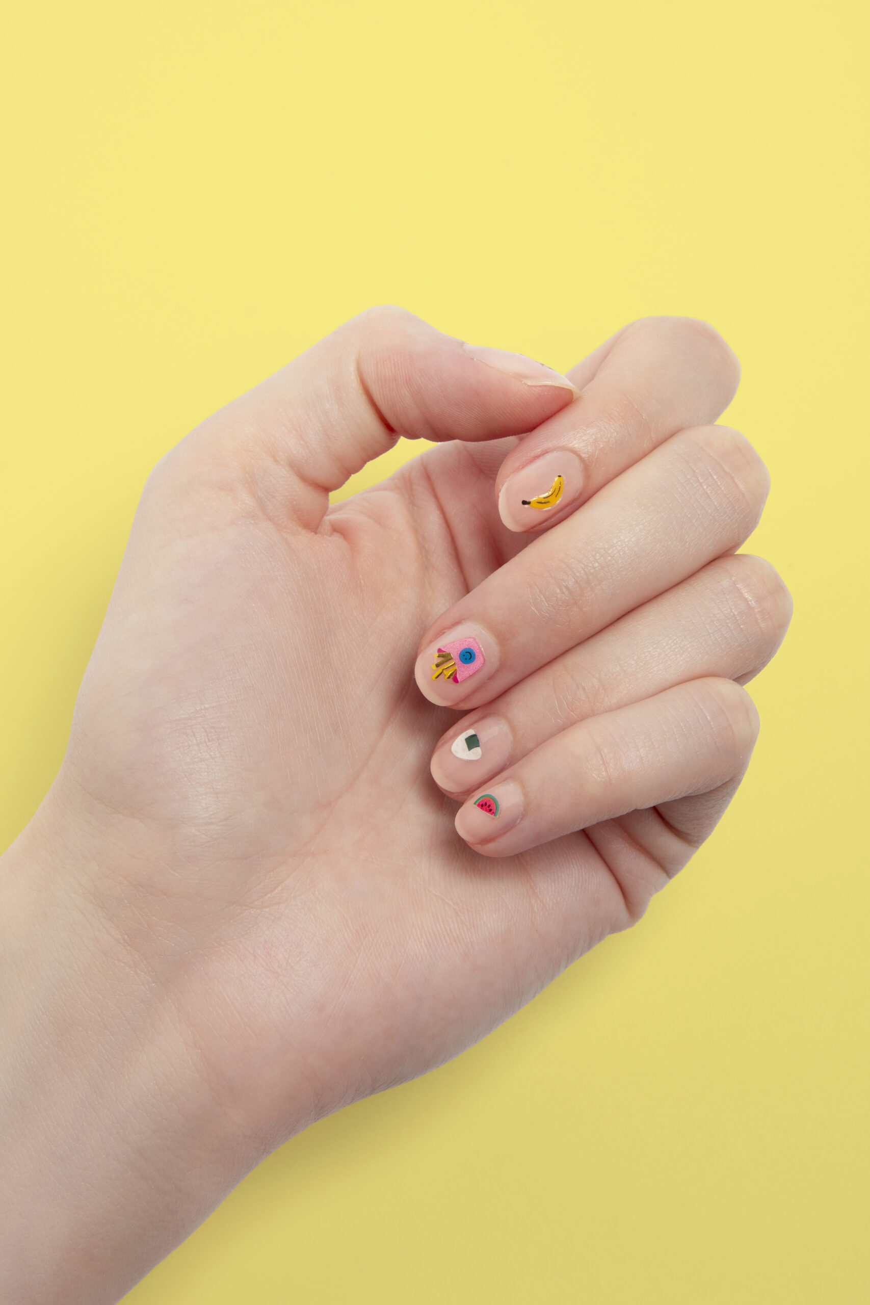 Main présentant une manucure colorée avec des motifs de banane, pastèque et sucette sur fond jaune