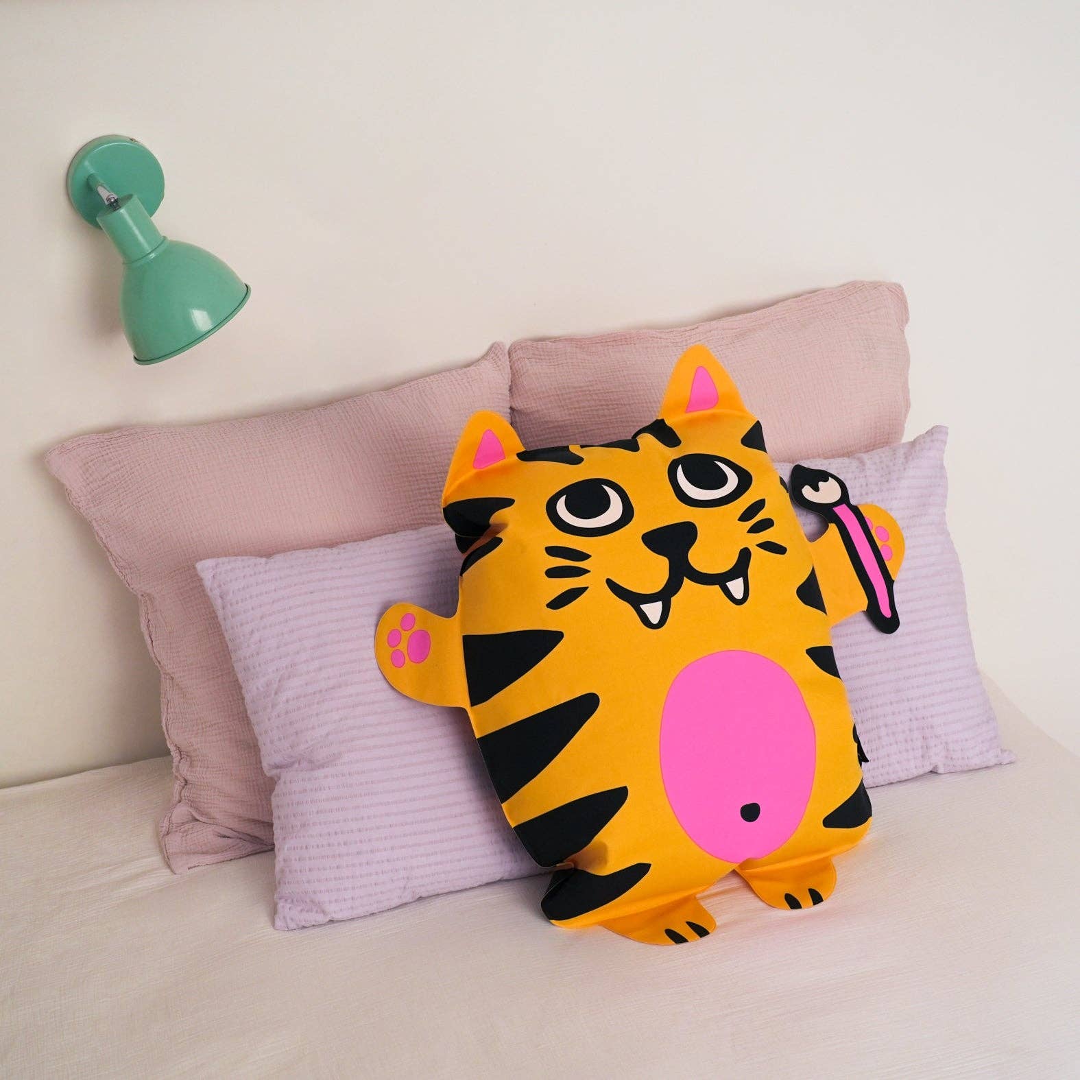 Coussin en forme de tigre orange posé sur des oreillers roses, avec une lampe murale verte